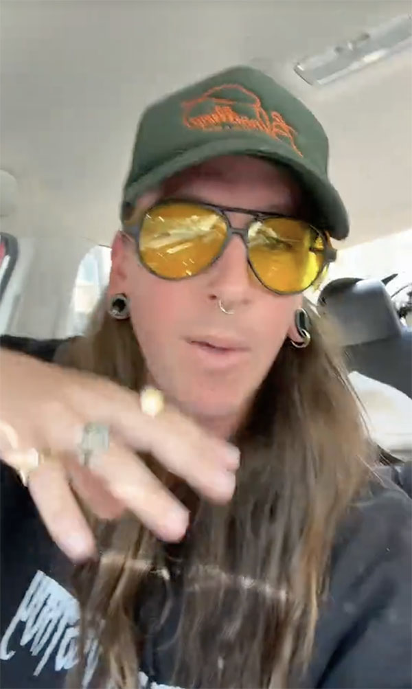 @acidblais aux longs cheveux bruns, lunettes de soleil jaunes et casquette verte, gesticulant dans un v&eacute;hicule